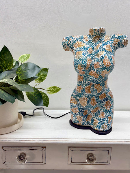 A ceramic torso-shaped lamp or figurine with a turquoise, white, and tan mosaic pattern sits on a white wooden cabinet next to a potted green plant.