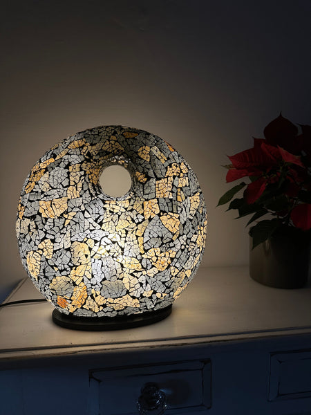 Handmade black and gold mosaic donut lamp illuminated with warm white LED light, creating a soft ambient glow on a table surface