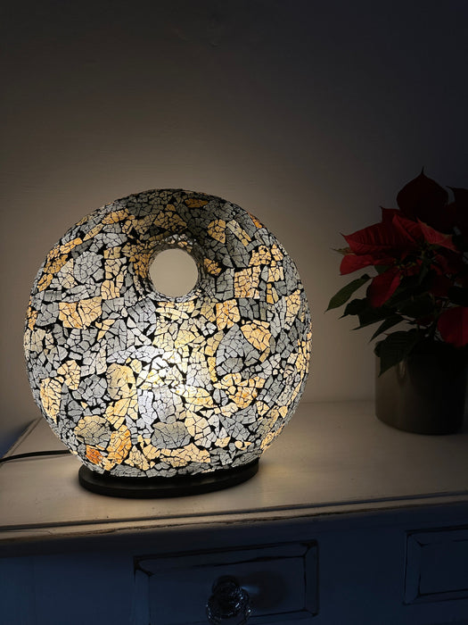 Handmade black and gold mosaic donut lamp illuminated with warm white LED light, creating a soft ambient glow on a table surface