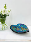 Handmade mosaic heart-shaped bowl in cyan tones on white table beside green flowers, showing blue and green glass pattern.