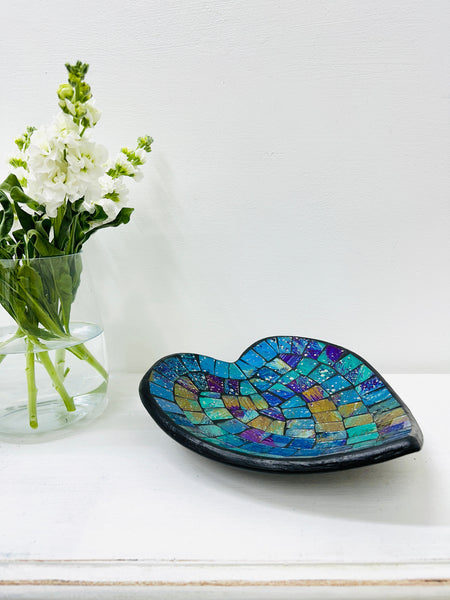 Handmade mosaic heart-shaped bowl in cyan tones on white table beside green flowers, showing blue and green glass pattern.