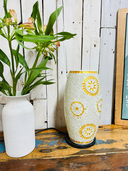 Handcrafted mosaic lamp in white and gold sunburst design, switched off beside a white vase with green plant on wooden surface.