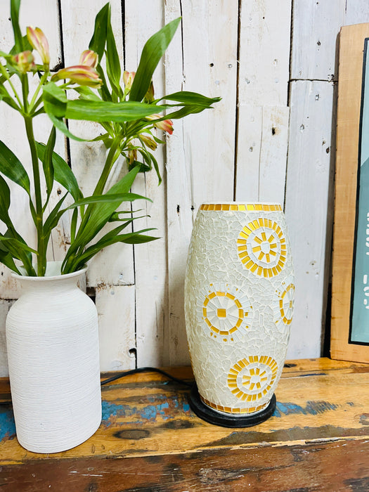 Handcrafted mosaic lamp in white and gold sunburst design, switched off beside a white vase with green plant on wooden surface.
