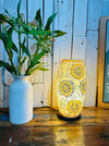 White and gold mosaic lamp with sunburst design beside green plant and vase, creating a warm decorative interior look.