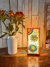Mosaic square lamp illuminated beside green plant in white vase on wooden table, giving warm ambient light.