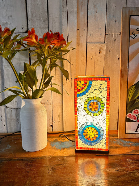 Mosaic square lamp illuminated beside green plant in white vase on wooden table, giving warm ambient light.