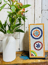 Handmade mosaic square lamp switched off on white wooden background beside green plant in white vase, showing colourful glass pattern and black cable.