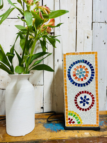 Handmade mosaic square lamp switched off on white wooden background beside green plant in white vase, showing colourful glass pattern and black cable.