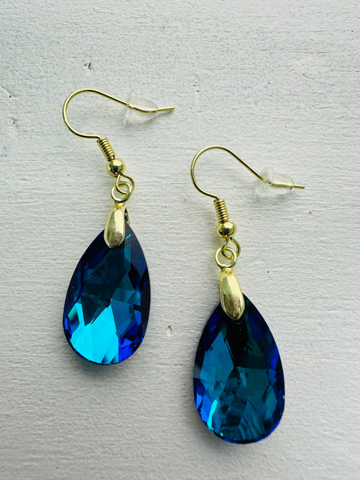 A pair of teardrop-shaped earrings with ocean blue crystals set in gold-colored settings, displayed against a white background.
