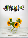 Colourful hand-painted metal parakeets on a branch displayed above a vase with sunflowers