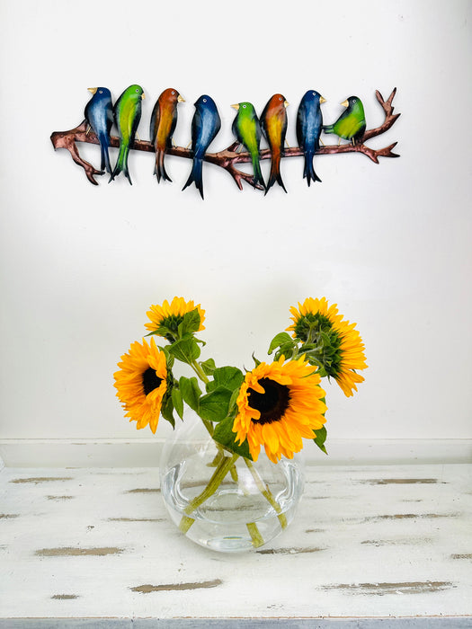 Colourful hand-painted metal parakeets on a branch displayed above a vase with sunflowers
