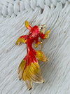 Goldfish-shaped brooch with red and yellow colours on a textured white background