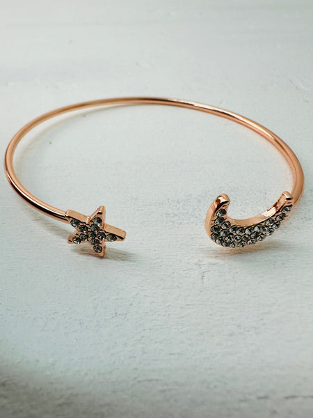 A rose gold cuff bracelet with a simple moon and star design, featuring black stones on the star and moon accents.