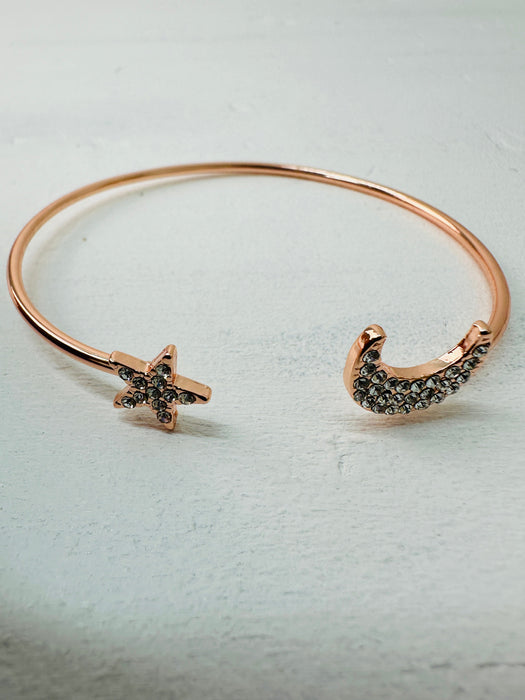 A rose gold cuff bracelet with a simple moon and star design, featuring black stones on the star and moon accents.