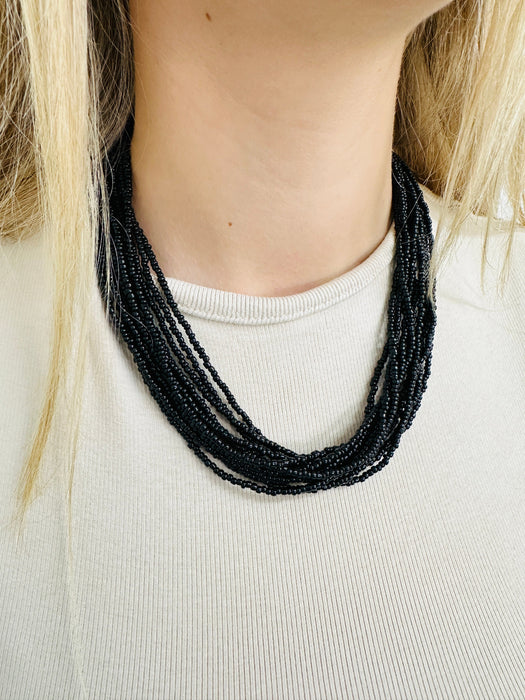 A person wearing a white top and showcasing a black necklace with multiple strands of seed beads.