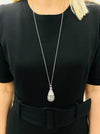 A person wearing a black dress with a silver-colored necklace featuring a teardrop-shaped pendant.