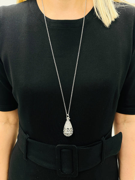A person wearing a black dress with a silver-colored necklace featuring a teardrop-shaped pendant.