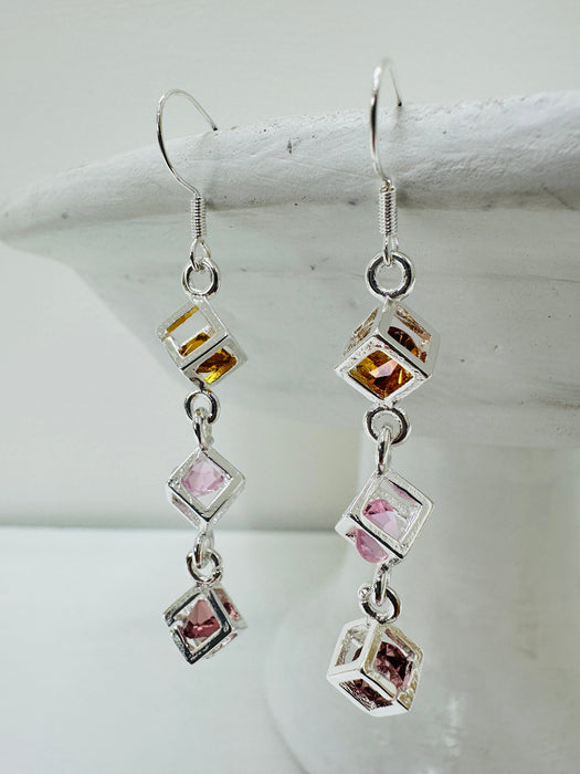 close up of earrings featuring triple crystal caged cubes with yellow, pink and purple crystal detail, displayed against a light background.