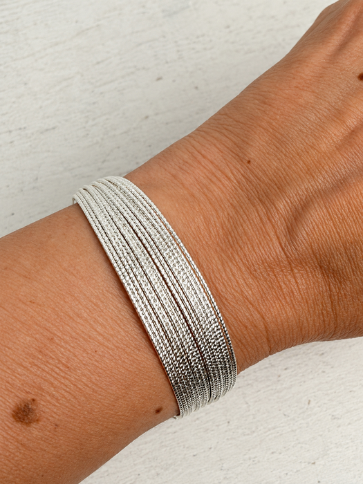 A close-up of a person's wrist wearing a multi-strand silver snake chain bracelet against a light textured background.
