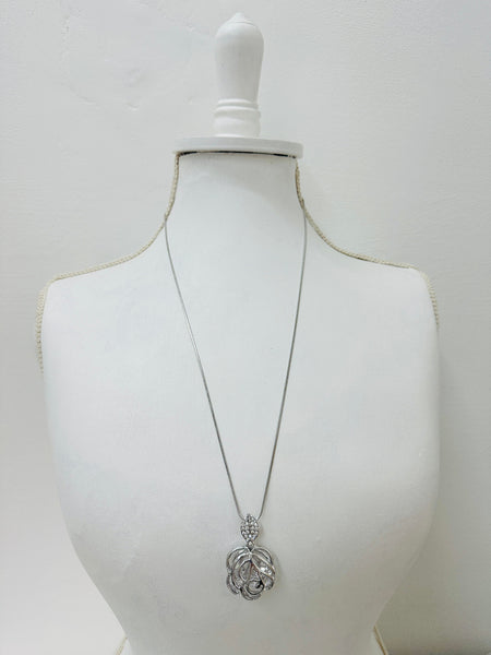 A silver-colored necklace with a long chain and a flower-style pendant, displayed on a mannequin bust.