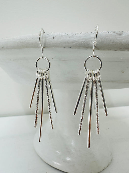 A pair of silver-colored earrings with a geometric design, featuring multiple dangling lines from a central circle.