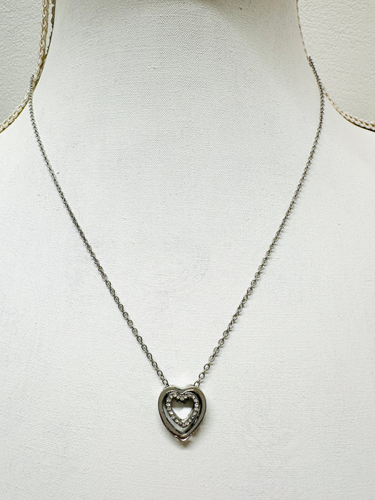 A silver necklace with a double heart pendant featuring a crystal detail, displayed on a mannequin.