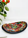 Small handmade mosaic oval bowl in Calypso tones on white table beside red flower, showing colourful glass mosaic pattern.