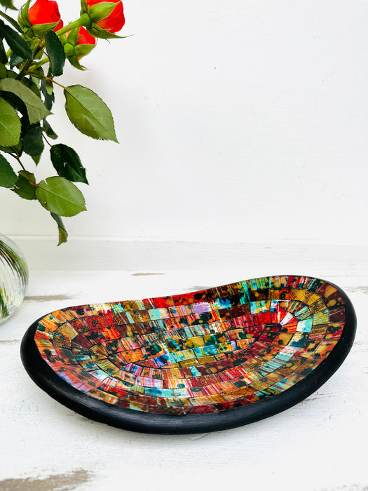 Small handmade mosaic oval bowl in Calypso tones on white table beside red flower, showing colourful glass mosaic pattern.
