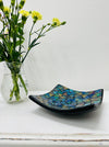 Handmade small mosaic square plate in cyan tones on white table beside yellow flowers, showing glossy blue and turquoise glass mosaic.