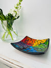 Handmade mosaic square dish in rainbow colours on white table beside green flowers, showing multicoloured glass mosaic and black rim.