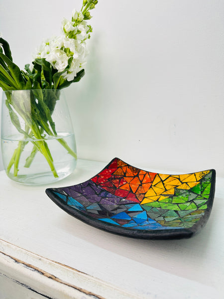 Handmade mosaic square dish in rainbow colours on white table beside green flowers, showing multicoloured glass mosaic and black rim.