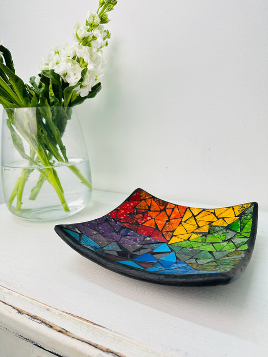 Handmade mosaic square dish in rainbow colours on white table beside green flowers, showing multicoloured glass mosaic and black rim.