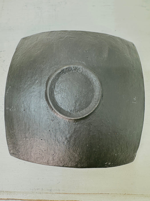 Underside of small mosaic square plate showing terracotta base and slightly curved edges on white surface.