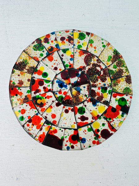 A circular mosaic coaster with a variety of colors splattered across its surface, with a hand-painted appearance.