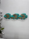 Three turquoise and brown fish-shaped wall hooks arranged in a row on a white wall, with a decorative white object and green leaves partially visible at the left edge.