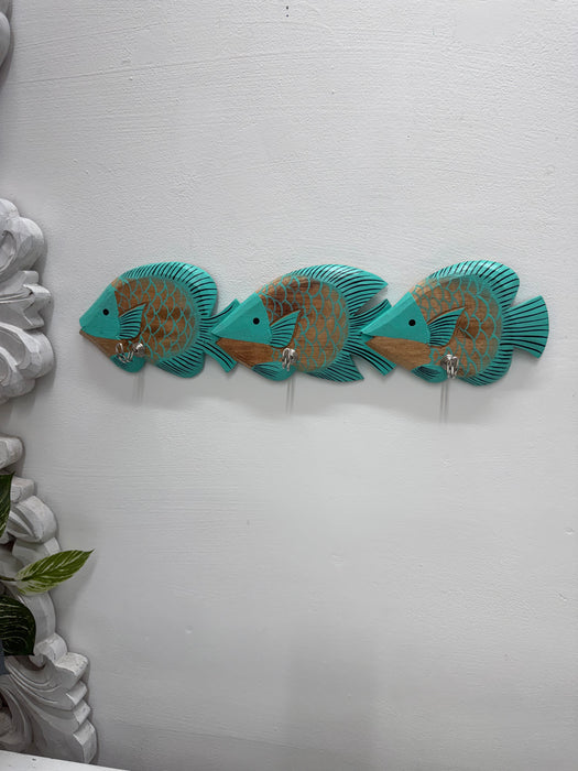 Three turquoise and brown fish-shaped wall hooks arranged in a row on a white wall, with a decorative white object and green leaves partially visible at the left edge.