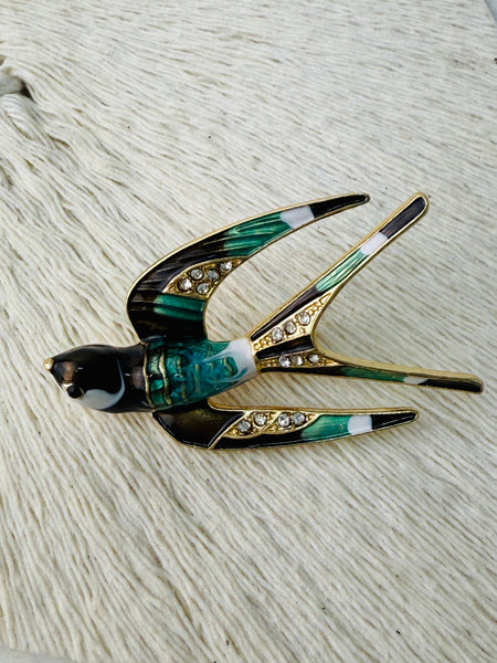 A brooch designed in the shape of a swallow bird with green and gold colors, featuring a black beak and white details on the bird's chest.