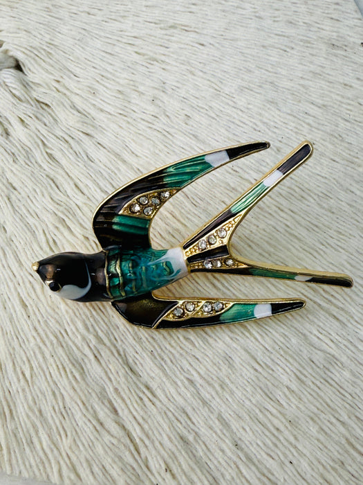 A brooch designed in the shape of a swallow bird with green and gold colors, featuring a black beak and white details on the bird's chest.