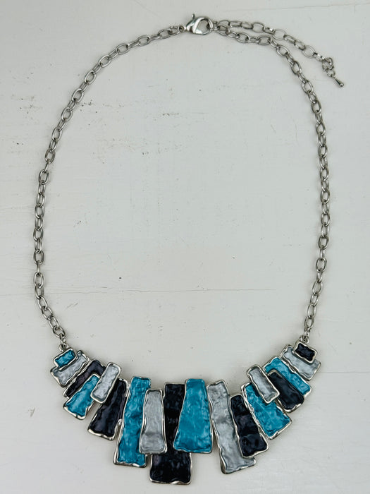 Blue enamel necklace with layered rectangular pieces in turquoise, grey and gunmetal, on a silver-tone chain, on a flatlay 