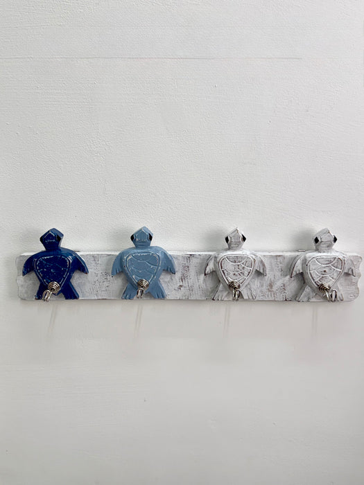 A rustic white-painted wooden wall hook rack featuring four turtle-shaped hooks in dark blue, light blue, and distressed white, mounted on a white wall.