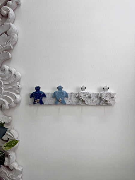 A decorative wall-mounted rack with four small turtle figurine-like hooks in blue, light blue, white, and gray, shown against a white wall with an ornate white frame on the left.