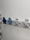 A decorative wall-mounted rack with four turtle-shaped hooks in blue and white, attached to a distressed white wooden bar on a white wall.