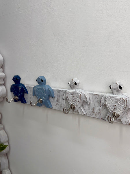 A decorative wall-mounted rack with four turtle-shaped hooks in blue and white, attached to a distressed white wooden bar on a white wall.