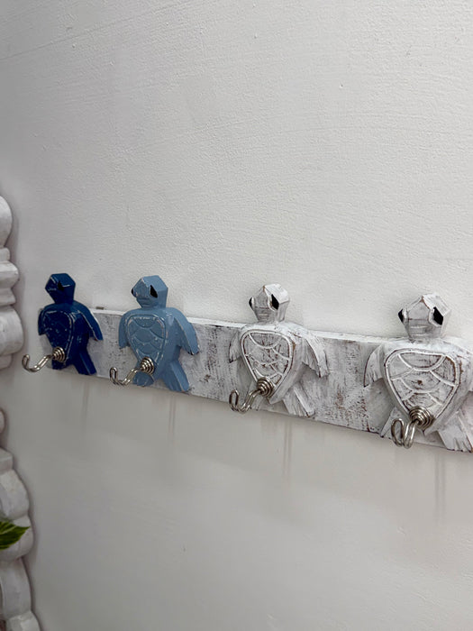 A decorative wall-mounted rack with four turtle-shaped hooks in blue and white, attached to a distressed white wooden bar on a white wall.