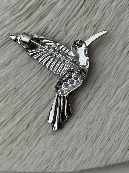 Back view of vibrant hummingbird brooch showing silver-tone metal pin with secure clasp on white textured background