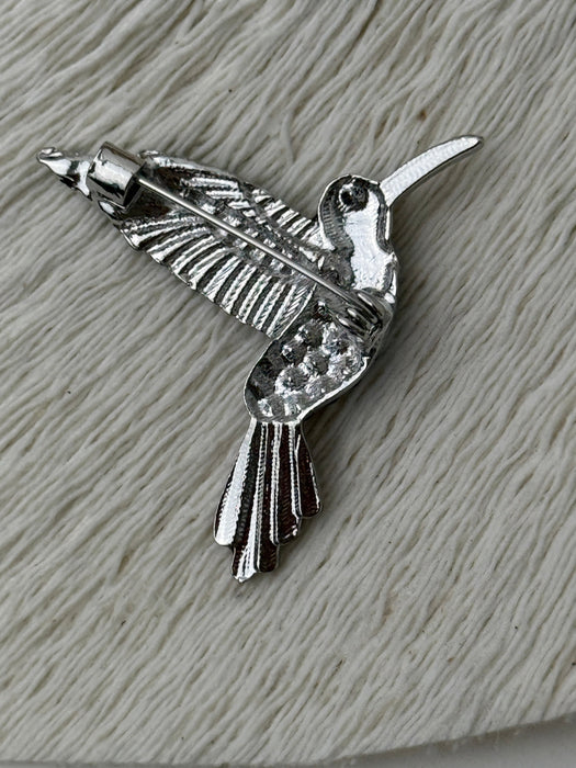Back view of vibrant hummingbird brooch showing silver-tone metal pin with secure clasp on white textured background