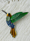 Decorative hummingbird brooch with green, blue, and bronze colours on a textured white background