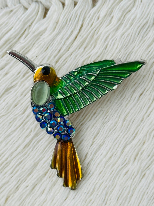 Decorative hummingbird brooch with green, blue, and bronze colours on a textured white background
