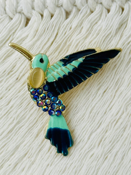 Hummingbird-shaped brooch with turquoise and dark turquoise colours on a textured white background