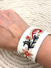 White bracelet with floral embroidery on a wrist against a textured white background